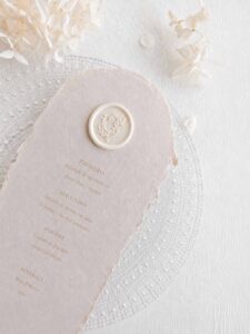 Sello de lacre adhesivo con diseño de guirnalda clásica colocado sobre un menú de boda de papel artesanal.