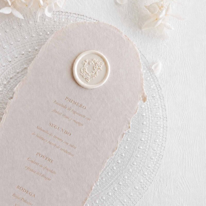 Sello de lacre adhesivo con diseño de guirnalda clásica colocado sobre un menú de boda de papel artesanal.