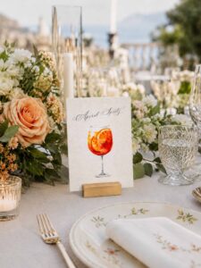 Número de mesa de boda Aperol Spritz en papel artesanal A6 con ilustración en acuarela, colocado sobre una mesa decorada con flores y velas.