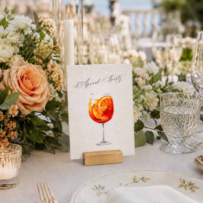Número de mesa de boda Aperol Spritz en papel artesanal A6 con ilustración en acuarela, colocado sobre una mesa decorada con flores y velas.