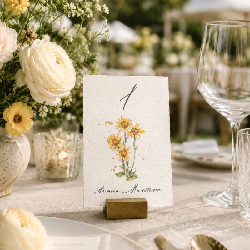Número de mesa de boda con ilustración de árnica en acuarela, impreso sobre papel artesanal y colocado en una mesa decorada con flores.