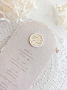 Sello de lacre adhesivo con diseño de flor dalia colocado sobre un menú de boda de papel artesanal.
