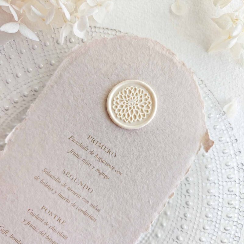 Sello de lacre adhesivo con diseño de flor dalia colocado sobre un menú de boda de papel artesanal.
