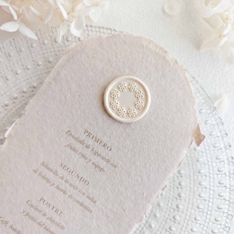 Sello de lacre adhesivo con diseño de guirnalda de flores colocado sobre un menú de boda de papel artesanal.