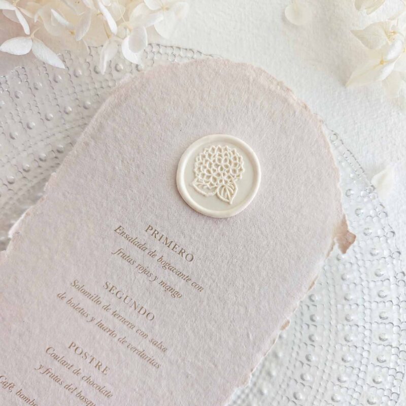 Sello de lacre adhesivo con diseño de hortensia colocado sobre un menú de boda de papel artesanal.