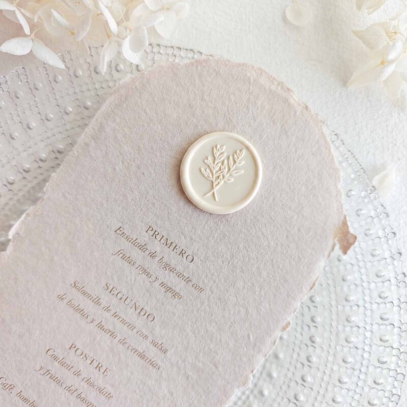 Sello de lacre adhesivo con diseño clásico de ramas cruzadas colocado sobre un menú de boda de papel artesanal.
