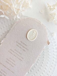 Sello de lacre adhesivo ovalado con diseño de clavel en 3D colocado sobre un menú de boda de papel artesanal.