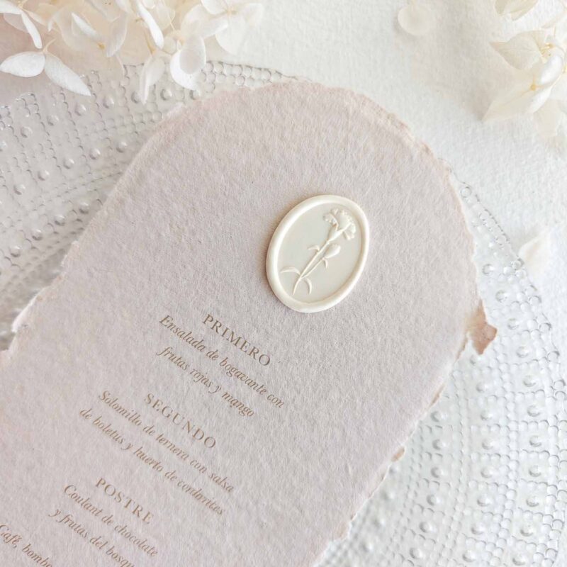 Sello de lacre adhesivo ovalado con diseño de clavel en 3D colocado sobre un menú de boda de papel artesanal.