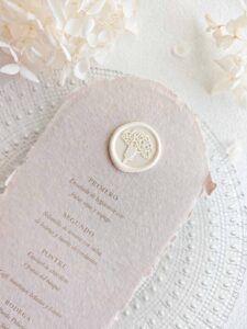 Sello de lacre adhesivo con diseño de flor clavel colocado sobre un menú de boda de papel artesanal.