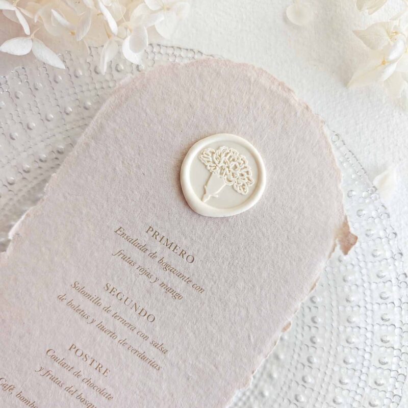 Sello de lacre adhesivo con diseño de flor clavel colocado sobre un menú de boda de papel artesanal.