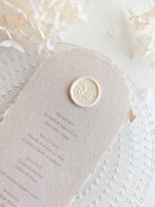 Sello de lacre adhesivo con diseño de flores en 3D colocado sobre un menú de boda de papel artesanal.