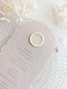 Sello de lacre adhesivo con diseño de olivo en 3D colocado sobre un menú de boda de papel artesanal.