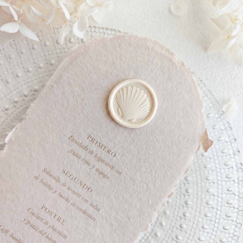 Sello de lacre adhesivo con diseño de concha en 3D colocado sobre un menú de boda de papel artesanal. Título de imagen