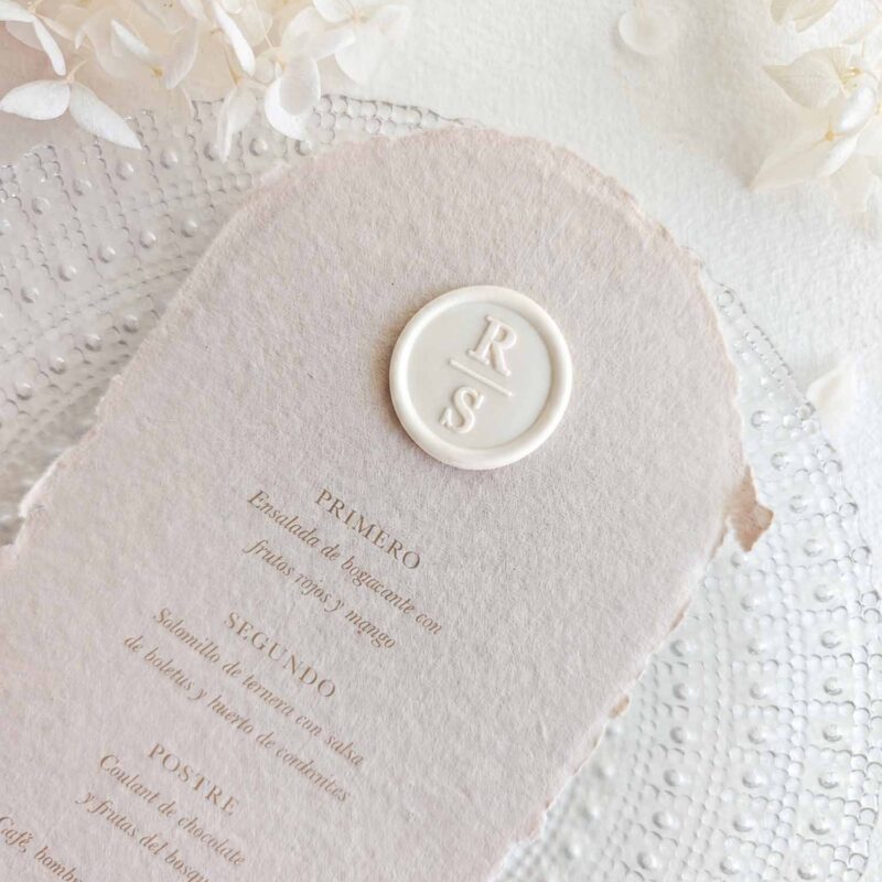 Sello de lacre adhesivo marfil personalizado con iniciales colocado sobre menú de boda artesanal.