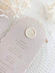 Sello de lacre adhesivo personalizado de estilo minimalista colocado sobre un menú de boda de papel artesanal.