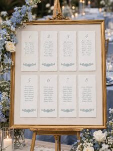 Seating plan de boda en papel artesanal cream con tarjetas individuales, marco dorado y decoración floral en tonos azul suave y blanco.