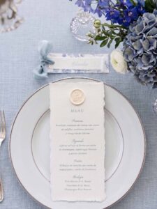 Menú de boda en papel artesanal Chloé con lacre personalizado sobre plato y puesto de mesa con cinta azul empolvado.