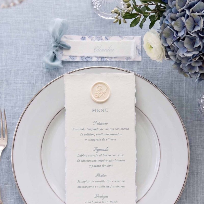 Menú de boda en papel artesanal Chloé con lacre personalizado sobre plato y puesto de mesa con cinta azul empolvado.