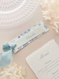 Puesto de mesa de boda Chloé con nombre del invitado en papel artesanal, estampado de hortensias y cinta azul empolvado.