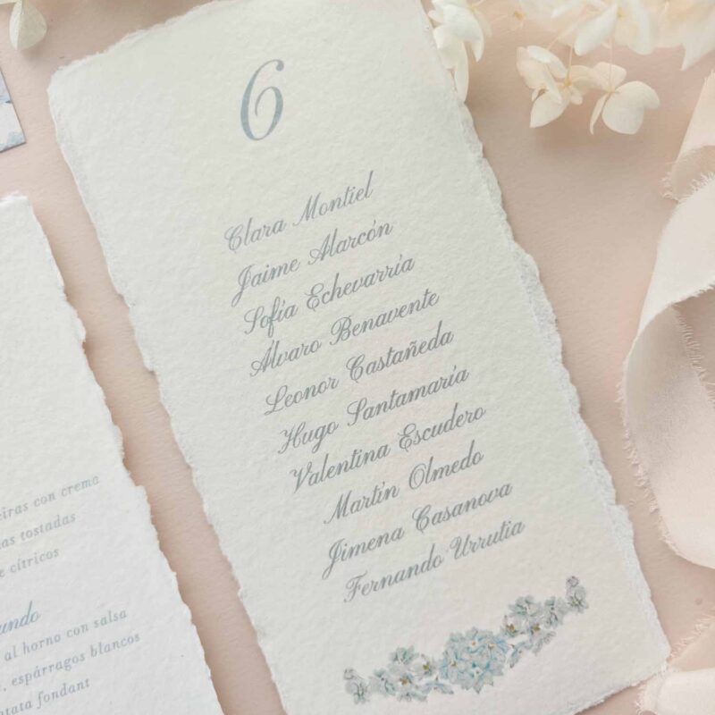 Seating plan Chloé en papel artesanal marfil con número de mesa y nombres de invitados impresos en azul empolvado.